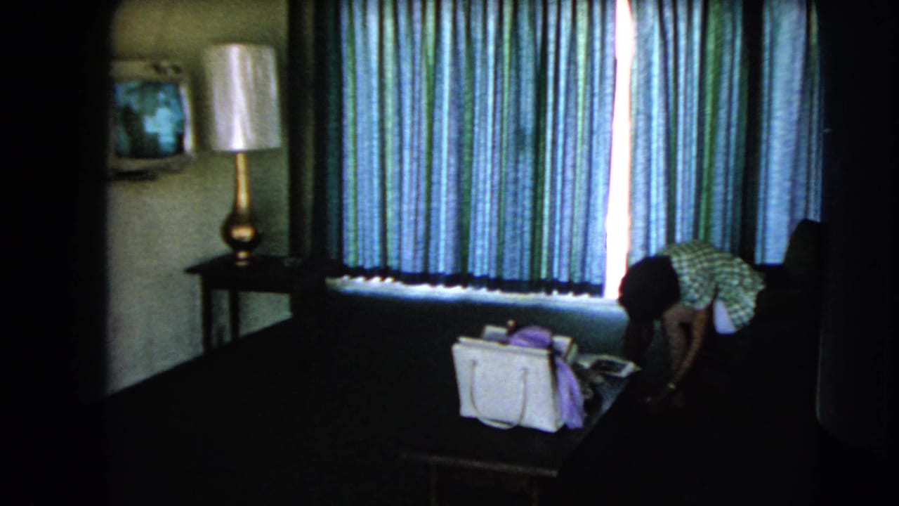 Woman bends near a table with a white handbag in a curtained hotel room.
