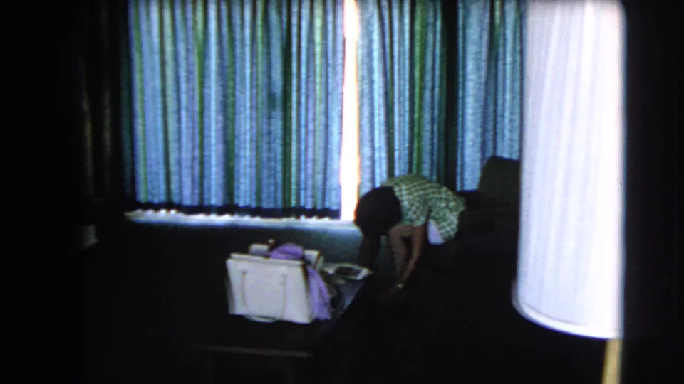 In the Las Vegas hotel room, a woman bends near a handbag while striped curtains and a large white lamp shade fill the frame.