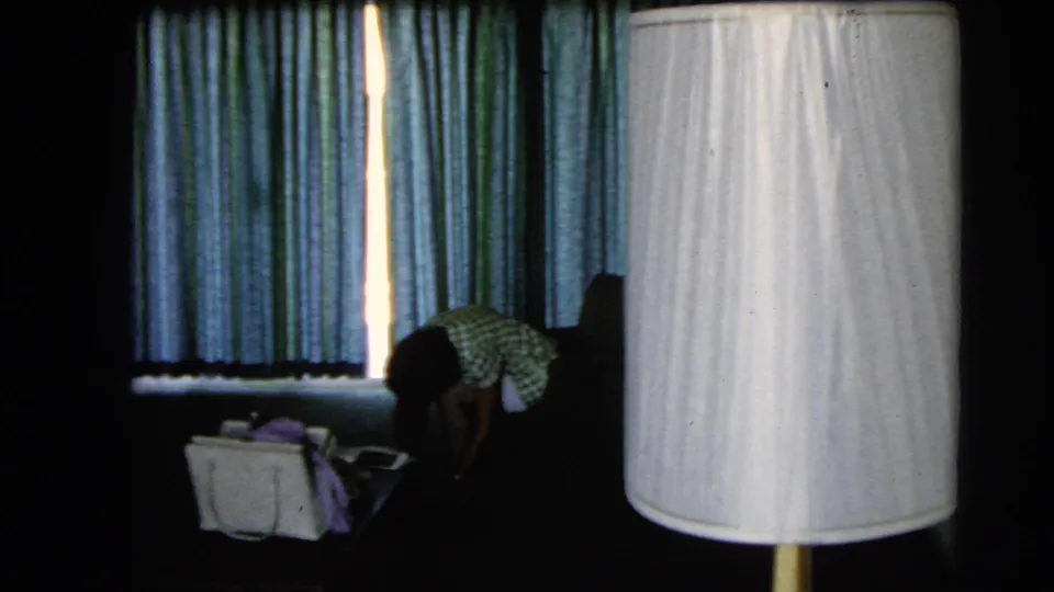 Large white lamp shade dominates the foreground as a woman bends near a handbag in the 1966 Las Vegas hotel room.