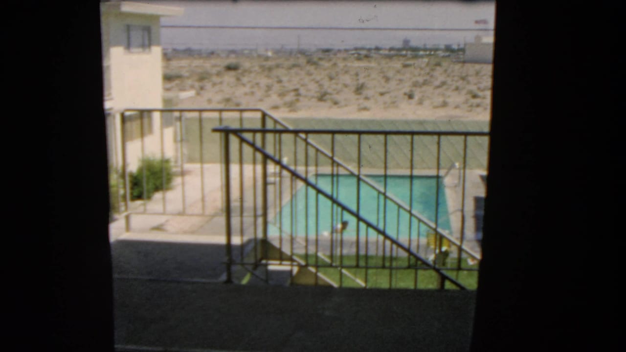 Balcony railing overlooks a turquoise swimming pool, pale building, and desert landscape beyond.