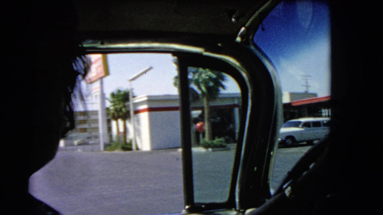 1966 Las Vegas home-movie frame seen from inside a car, with a dark passenger silhouette, window frame, roadside gas station, palm trees, and parked car visible outside.