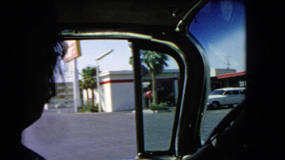 1966 Las Vegas home-movie frame seen from inside a car, with a dark passenger silhouette, window frame, roadside gas station, palm trees, and parked car visible outside.