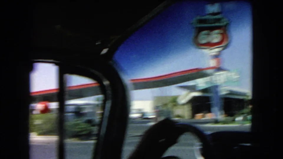 From inside the moving car, a Route 66-style gas station sign, red-trimmed roadside building, steering wheel silhouette, parked cars, and bright sky pass by in 1966 Las Vegas footage.