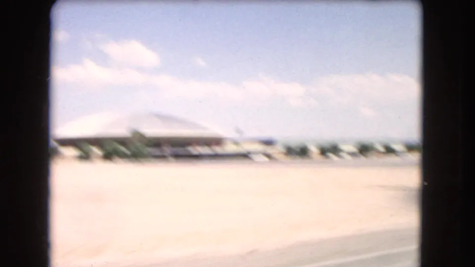 A blurred 8mm frame through the car window shows bright open pavement, low roadside buildings, distant parked cars, and pale sky during the Las Vegas driving sequence.
