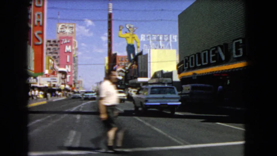1966 Las Vegas street frame looking down the boulevard with Golden Nugget signage, a Lucky figure sign, traffic, and a pedestrian crossing.