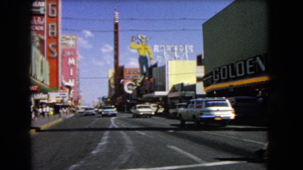 Moving street view with casino signs, classic cars, sidewalks, and a bright blue sky.