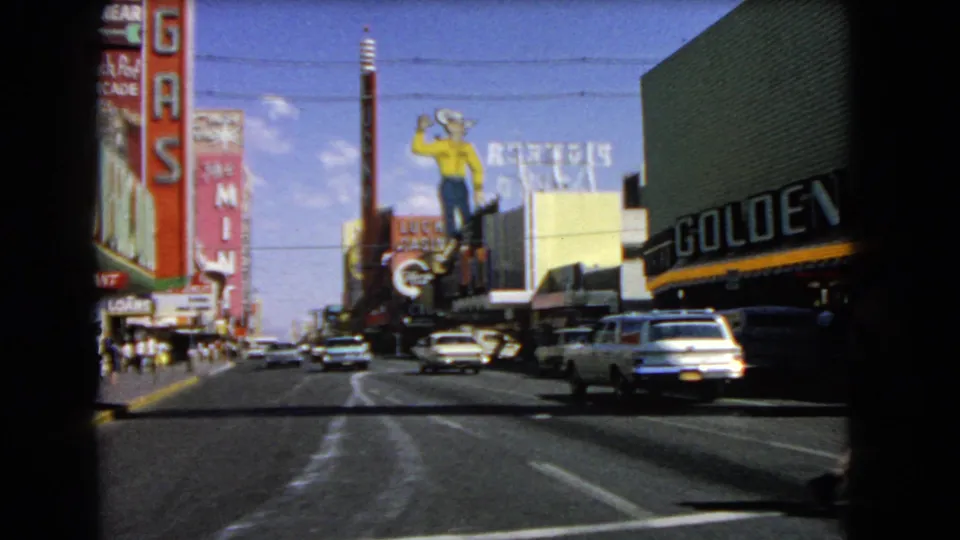 1966 Las Vegas street view from a moving vehicle, with casino signage, mid-century cars, pedestrians, and overhead wires.