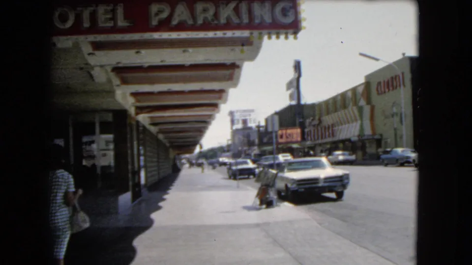 1966 Las Vegas street view with a hotel parking sign, casino or hotel storefronts, pedestrians, and mid-century cars in traffic.