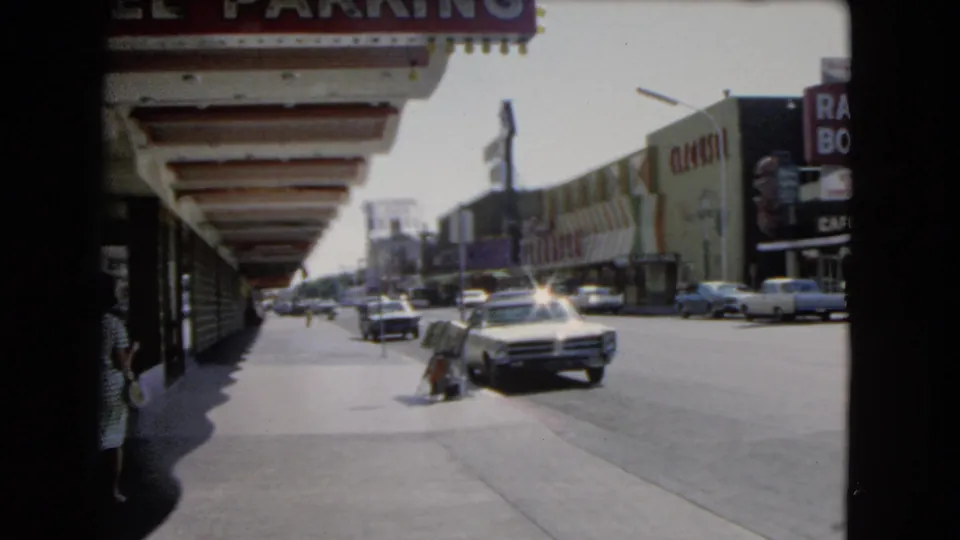 Las Vegas street footage from 1966 showing casino facades, a wide sidewalk, passing cars, and a pedestrian near the curb.