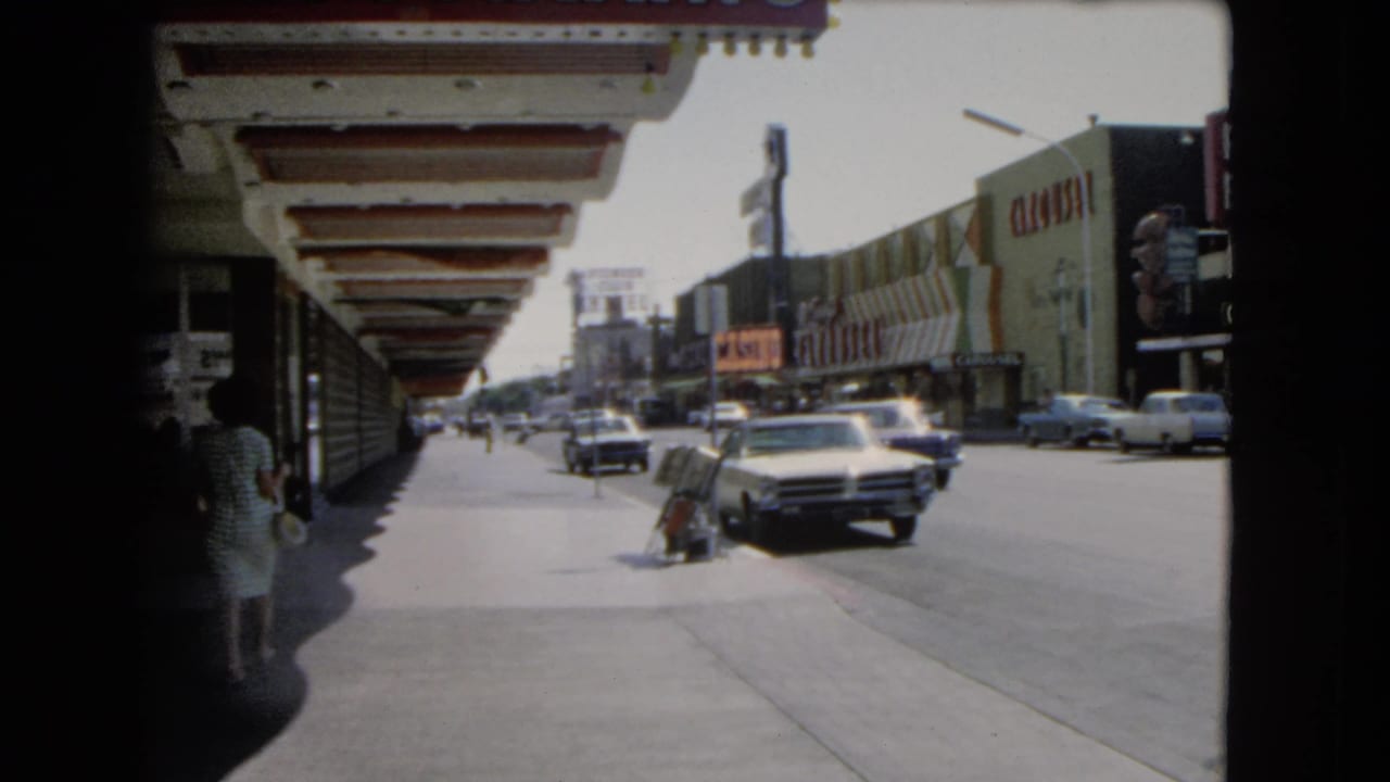 1966 Las Vegas downtown street view with classic cars, pedestrians, shaded sidewalk overhangs, and storefront signage.