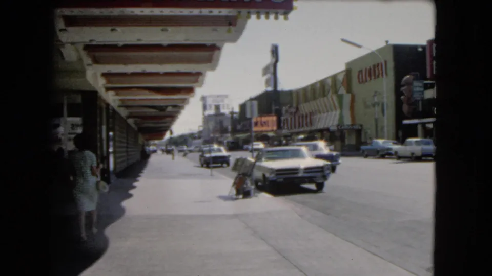 1966 Las Vegas downtown street view with classic cars, pedestrians, shaded sidewalk overhangs, and storefront signage.