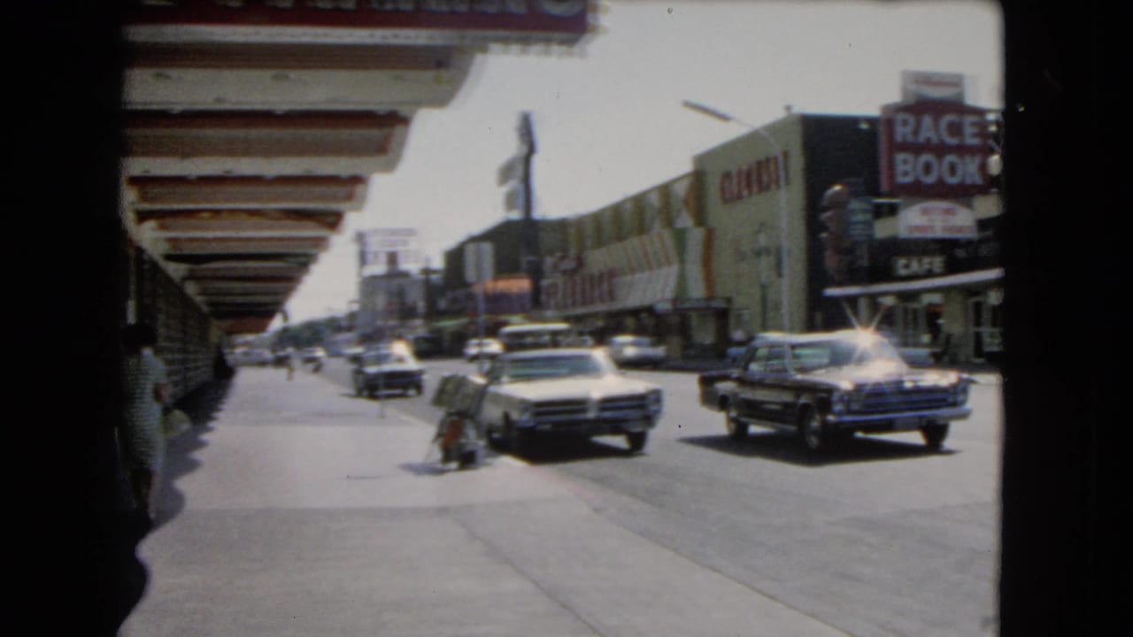 Classic cars pass downtown storefronts with a race book sign and pedestrians on the sidewalk.