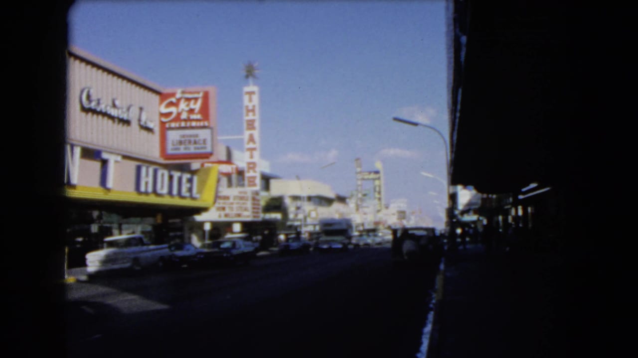 1966 Las Vegas street view showing Mint Hotel signage, a vertical theatre sign, moving cars, blue sky, and shaded framing.