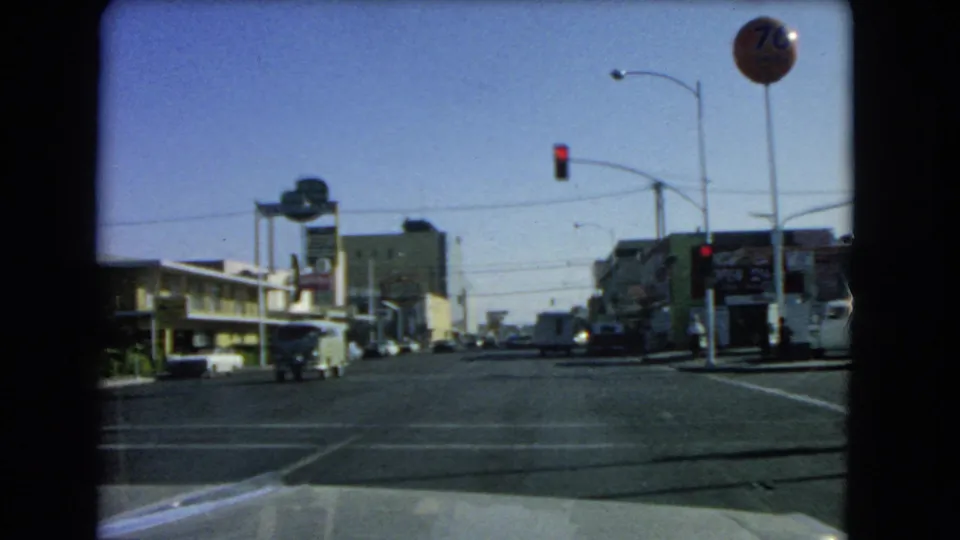 1966 Las Vegas windshield view at an intersection, with a red traffic signal, round roadside sign, classic cars, and commercial street frontage.