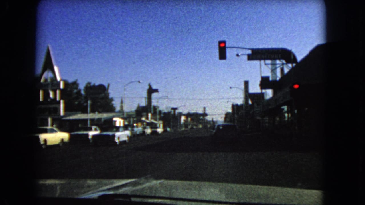Street traffic seen from a car, with a red signal overhead and roadside buildings in shadow.