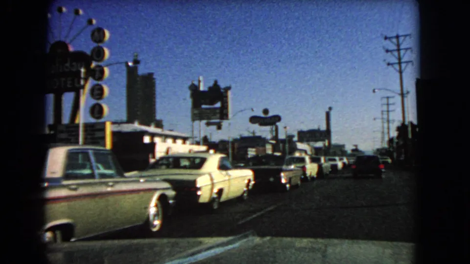 View from inside a car on a 1966 Las Vegas street, with mid-century automobiles, motel signs, utility poles, and dark rounded film-frame borders.
