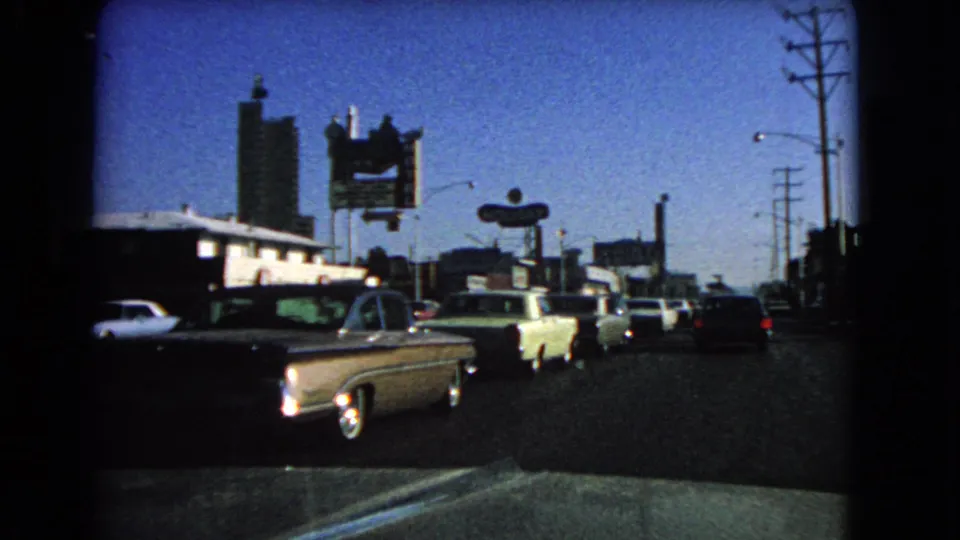 Close vehicle-side view of 1966 Las Vegas traffic, with mid-century cars, motel signage, utility poles, and commercial storefronts.