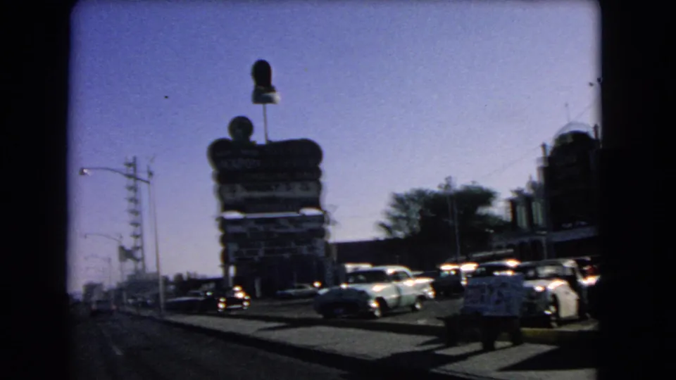 1966 Las Vegas roadside view with parked cars, a large Holiday-style sign, sidewalk, utility poles, and low commercial buildings.
