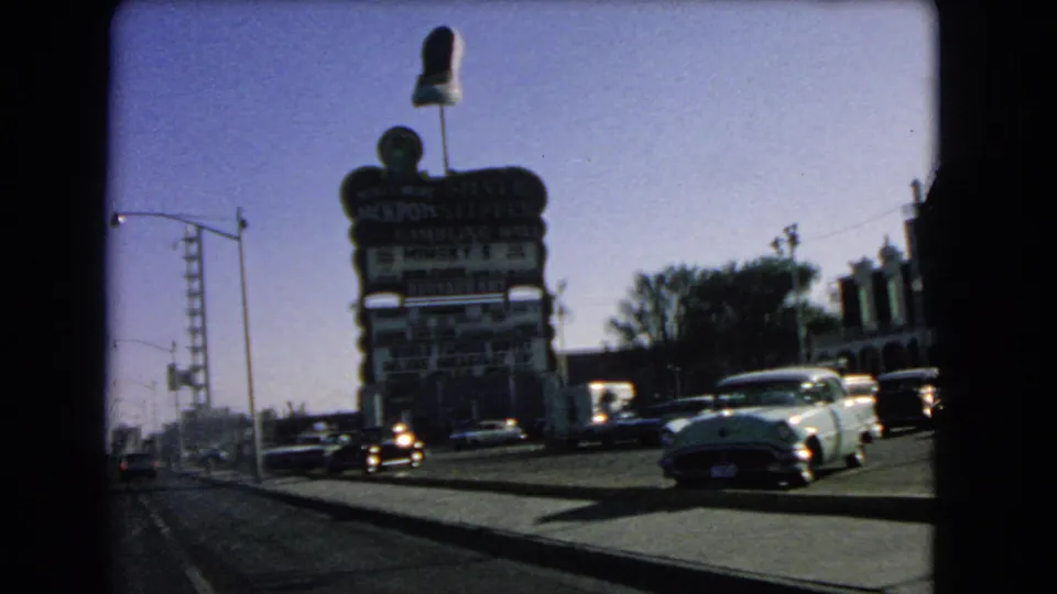 A 1966 Las Vegas roadway scene shows mid-century cars moving past a large entertainment sign and low commercial buildings.