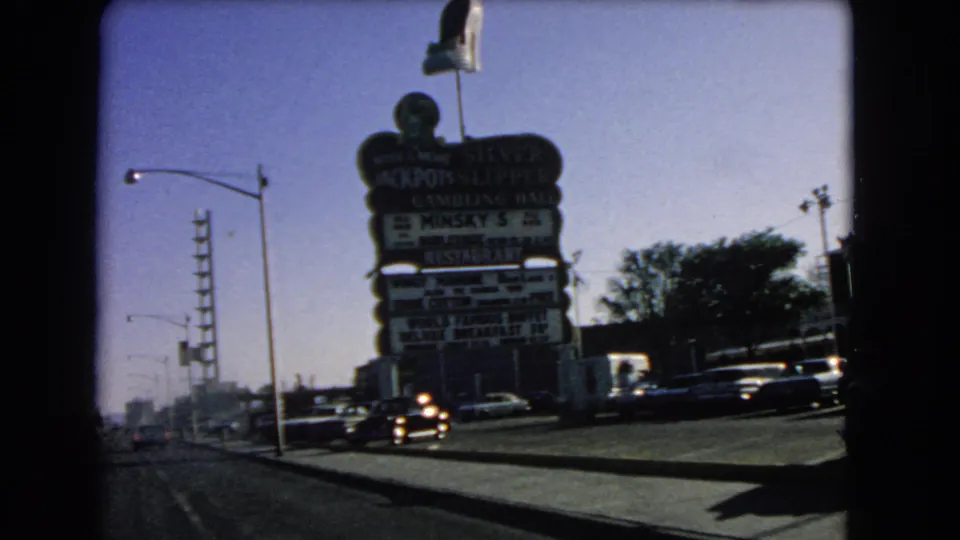 The vehicle draws closer to a Las Vegas gambling hall and restaurant sign, with 1960s cars, roadway poles, and sidewalk visible.