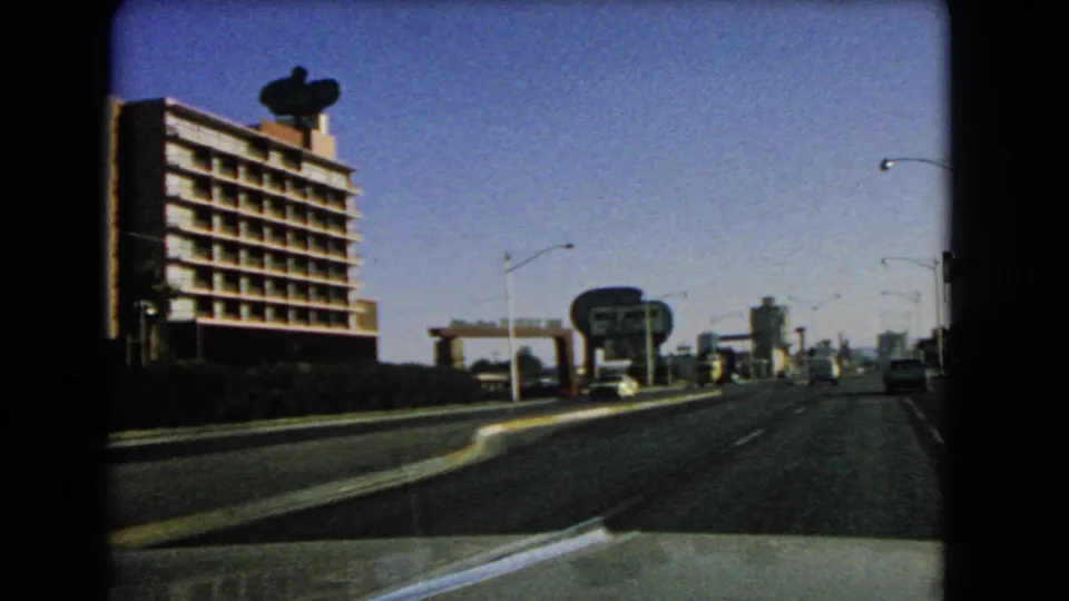 A 1966 Las Vegas driving view opens to a broader street, showing a multi-story building, rooftop sign, smaller roadside sign, and sparse traffic.