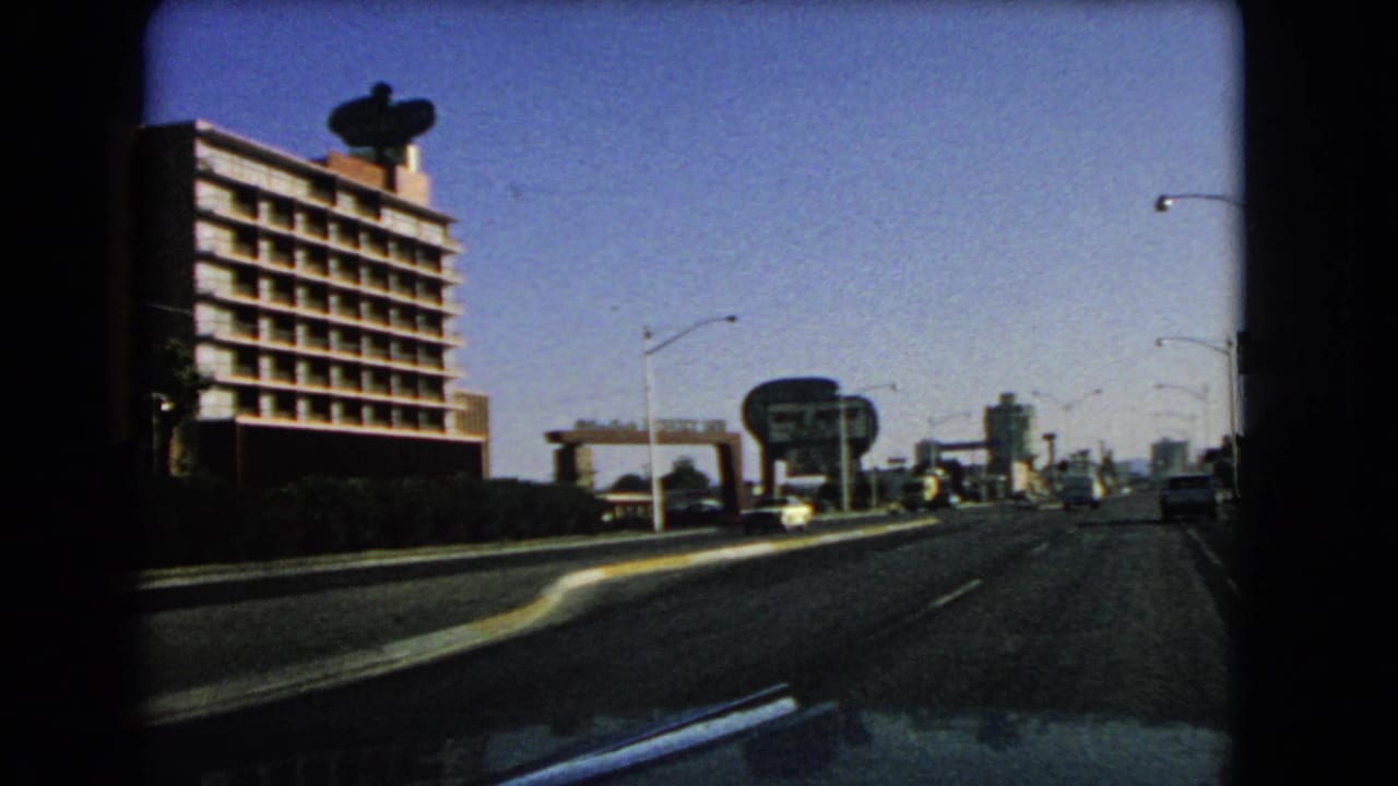 Roadway seen through a windshield with a multi-story building, rooftop dish-like shape, cars, and roadside signs.