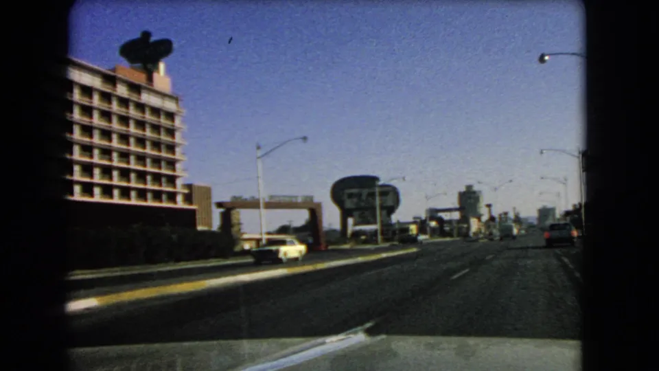 1966 Las Vegas street view with a light-colored car, multi-story roadside building, tall streetlights, large signs, and distant towers.