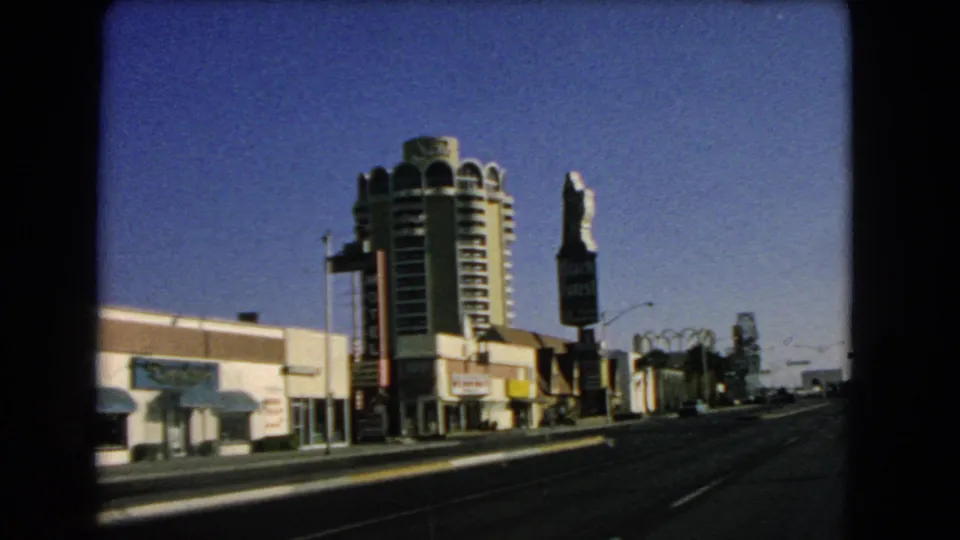 1966 Las Vegas roadway view showing a tall round building, low storefronts, vertical roadside signage, streetlights, and distant structures.