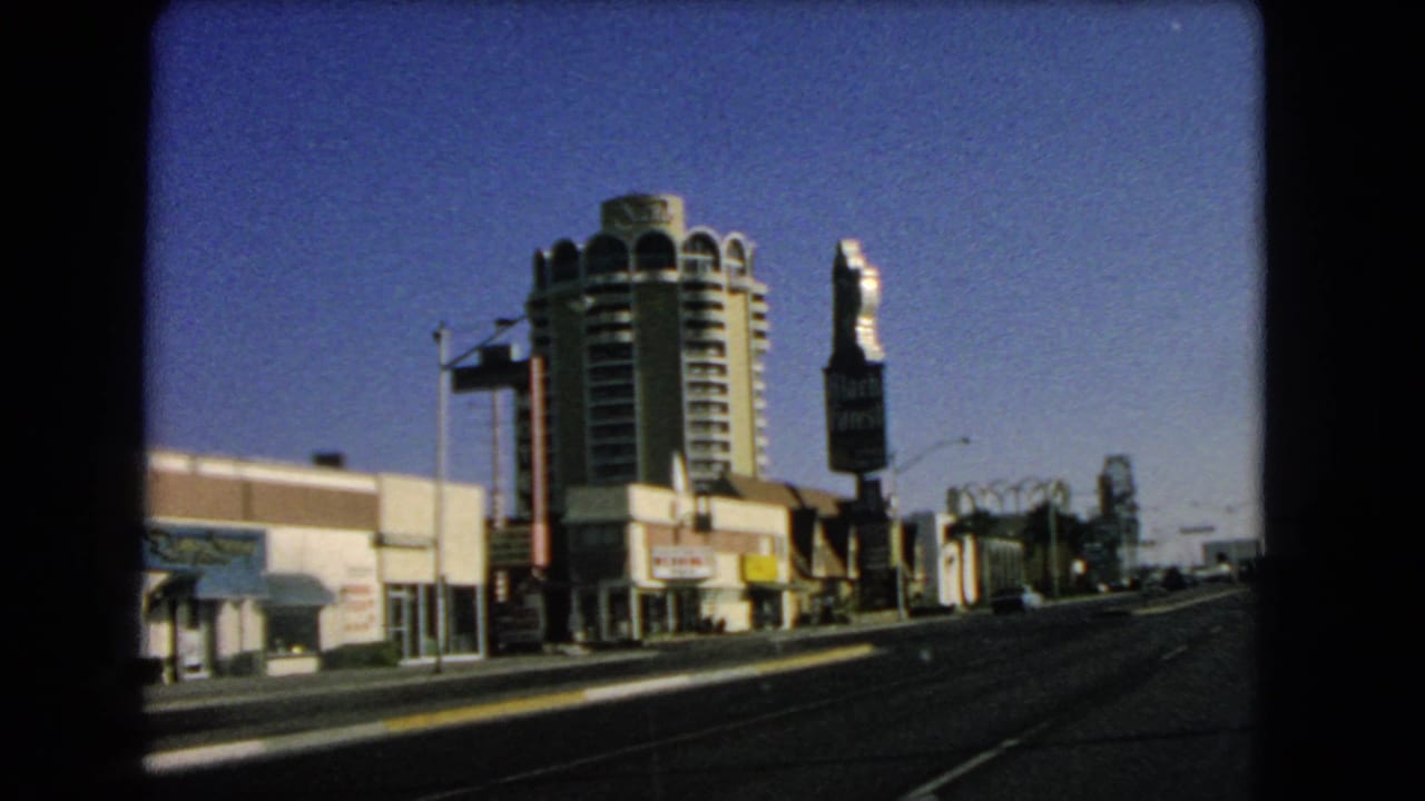 1966 Las Vegas street view with broad roadway, low commercial frontage, cylindrical hotel-like tower, vertical signage, and clear sky.