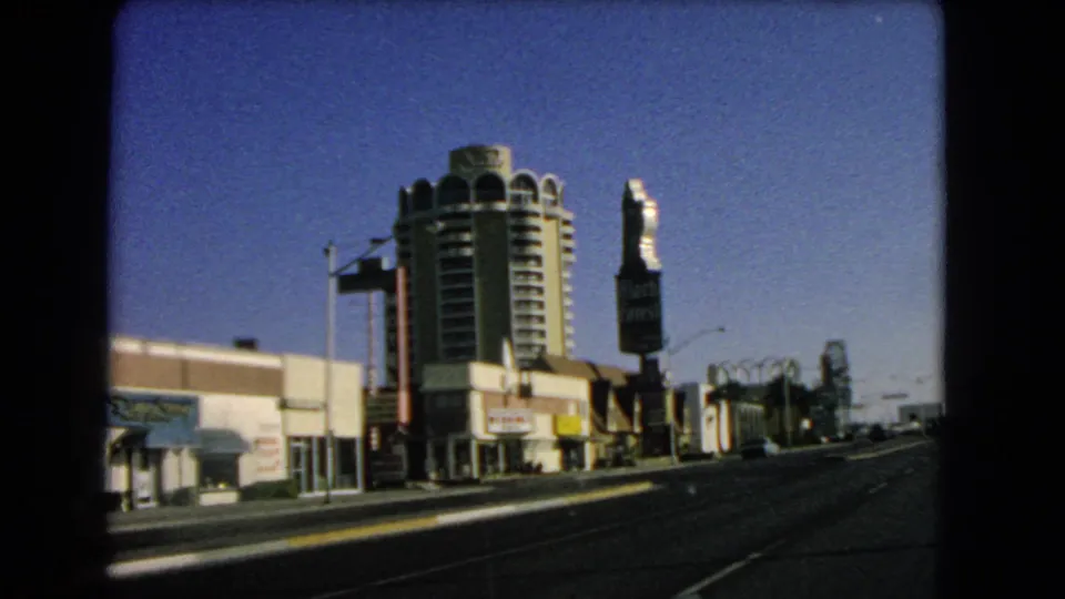 1966 Las Vegas street view with broad roadway, low commercial frontage, cylindrical hotel-like tower, vertical signage, and clear sky.