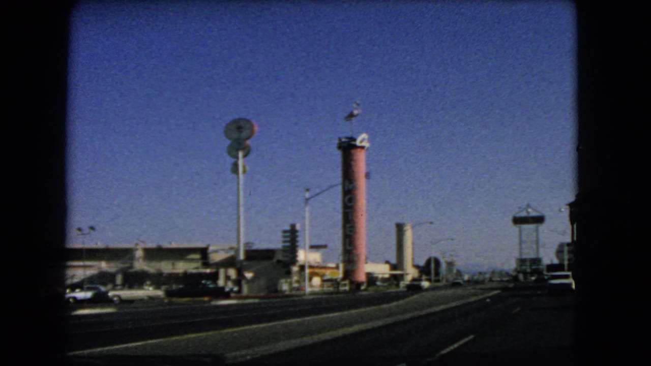 1966 Las Vegas roadside scene showing a tall pink sign, broad traffic lanes, parked cars, low buildings, and desert sky.