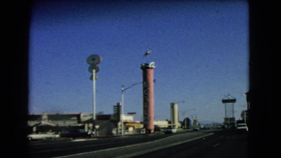 Las Vegas roadside frame from 1966 with vertical signage, streetlights, parked cars, low commercial structures, and broad asphalt lanes.