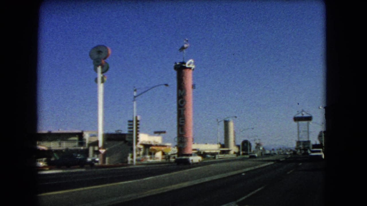 1966 Las Vegas drive-by view showing a tall pink cylindrical motel sign, roadside buildings, streetlights, and a clear blue sky.