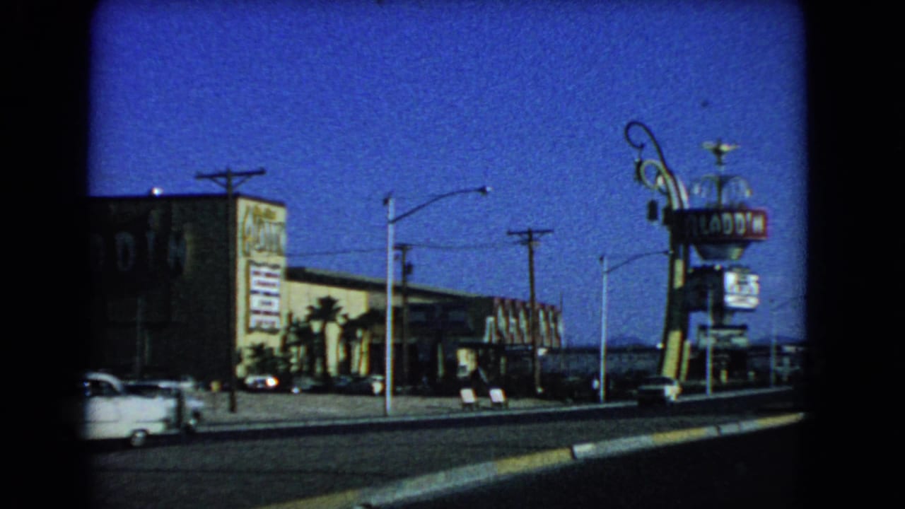 1966 Las Vegas roadside scene showing an Aladdin-themed sign, motel frontage, parked cars, palms, utility poles, and a bright blue sky.