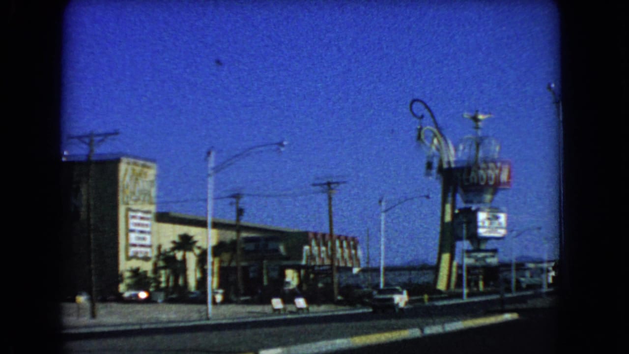 1966 Las Vegas drive-by frame with an Aladdin-themed roadside sign, low commercial buildings, parked cars, and tall streetlights.