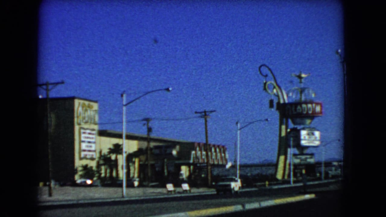 Street view of the Aladdin Hotel sign and exterior with cars and palm trees.