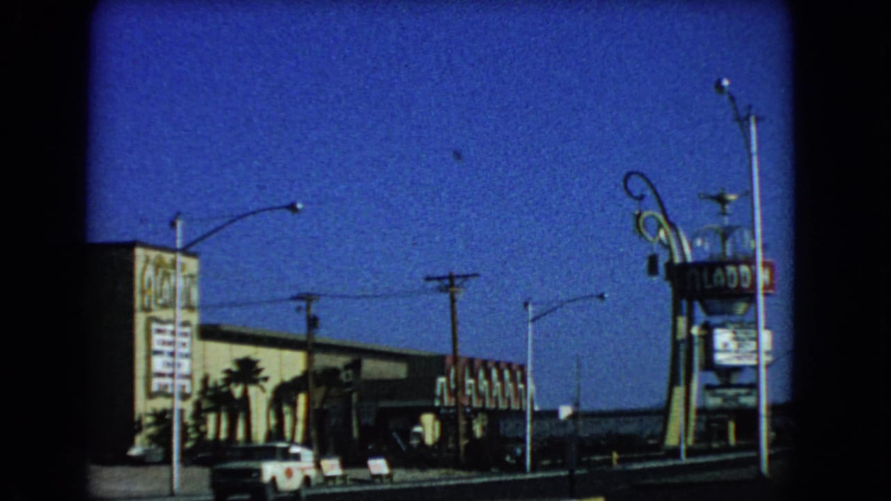 1966 Las Vegas street frame showing the Aladdin sign, hotel exterior, a white car, palm trees, signs, and overhead utility lines.