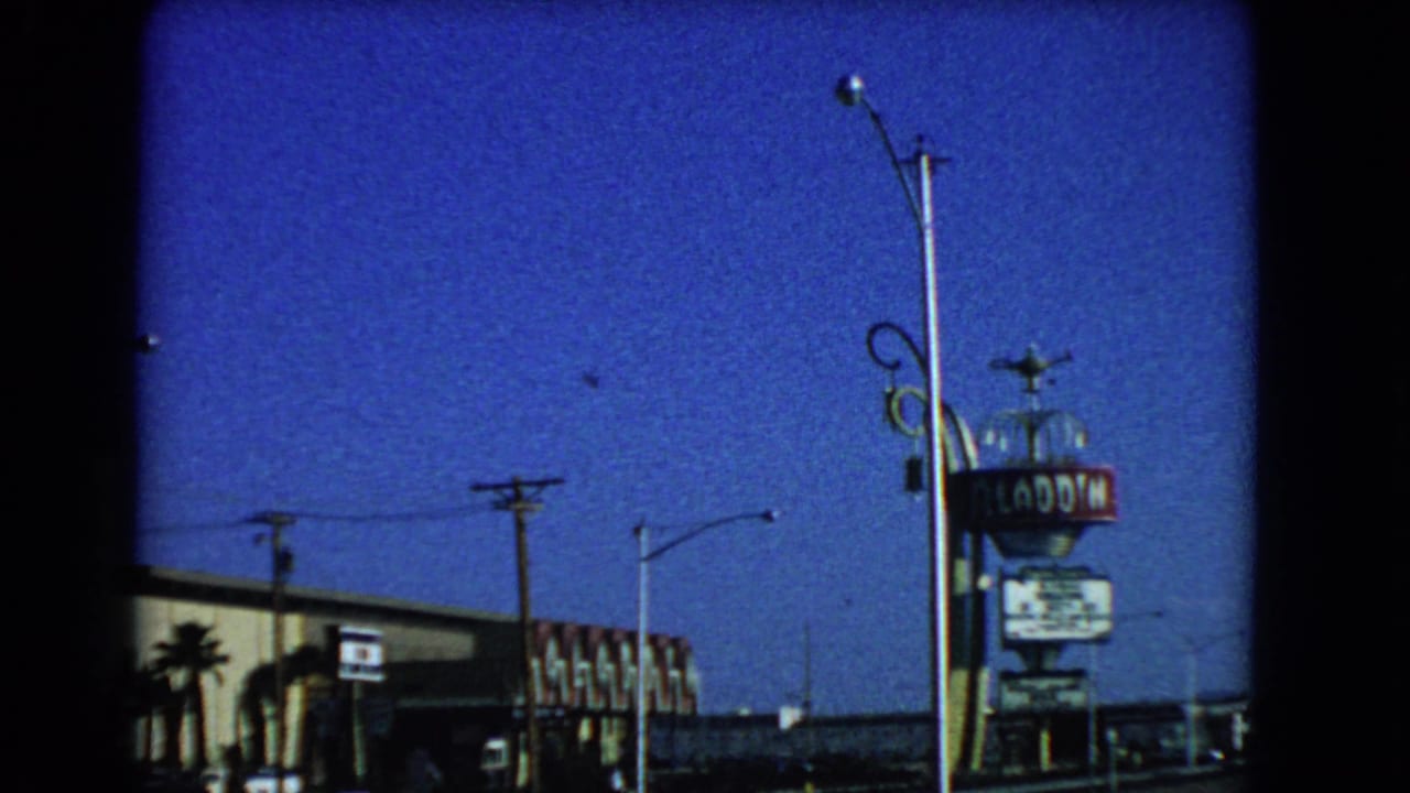 1966 Las Vegas 8mm frame showing the Aladdin Hotel sign, streetlights, utility poles, and roadside buildings under a blue sky.