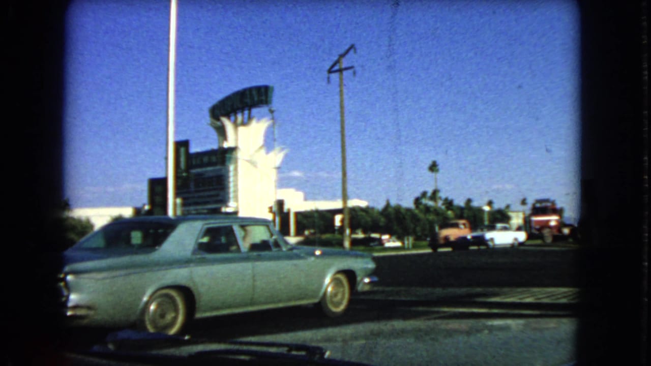 1966 Las Vegas street frame showing a light-colored classic sedan, hotel frontage, tall sign structure, traffic, and road markings.