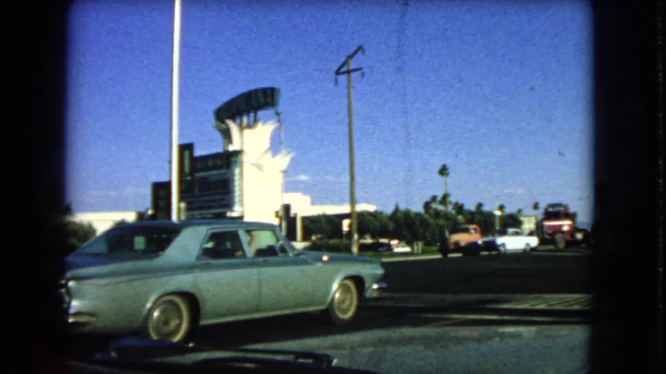 1966 Las Vegas street frame showing a light-colored classic sedan, hotel frontage, tall sign structure, traffic, and road markings.