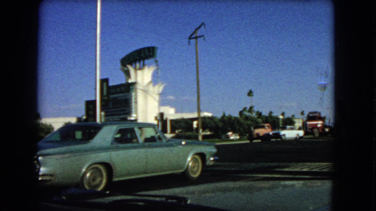 1966 Las Vegas roadside view with a pale blue sedan in the foreground, other vehicles beyond, palm trees and a white curved-roof building.