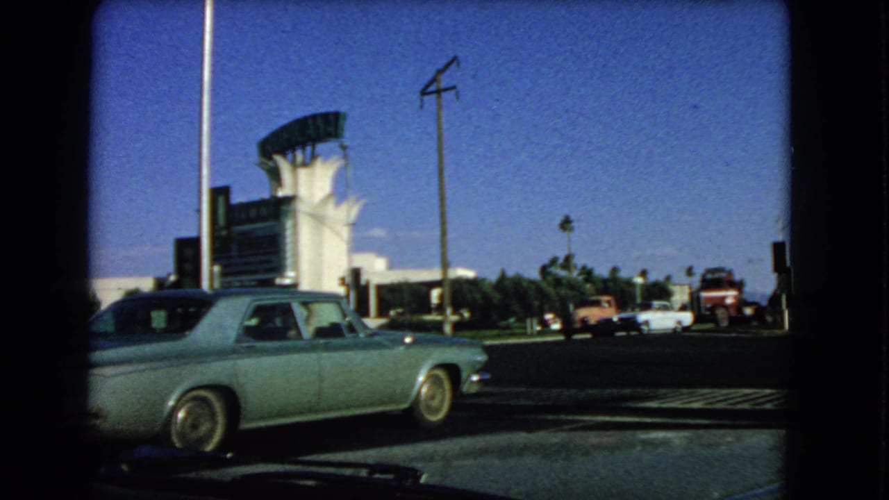 Vintage sedan in front of roadside Las Vegas signage, palm trees and parked vehicles.
