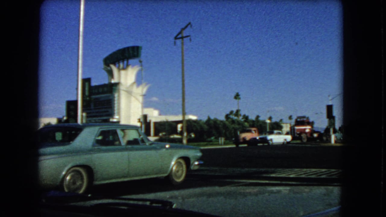 1966 Las Vegas home-movie frame showing a pale blue classic sedan, a large sign structure, white building and vintage vehicles along the street.