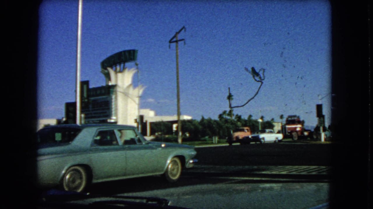 1966 Las Vegas frame with the foreground sedan partly leaving view, parked vehicles near a sign structure, palm trees and visible 8mm film marks.
