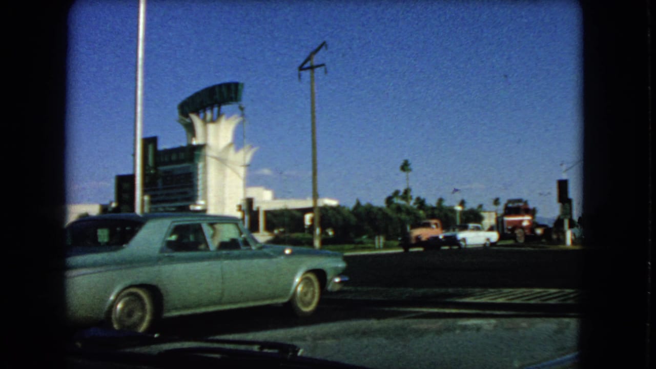 Vintage cars pass a white roadside building as a blue sedan remains in the foreground.