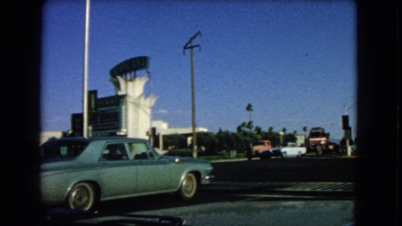 1966 Las Vegas home-movie frame with a blue sedan cropped at left, a distinctive white building, utility poles, palms, and vintage traffic.