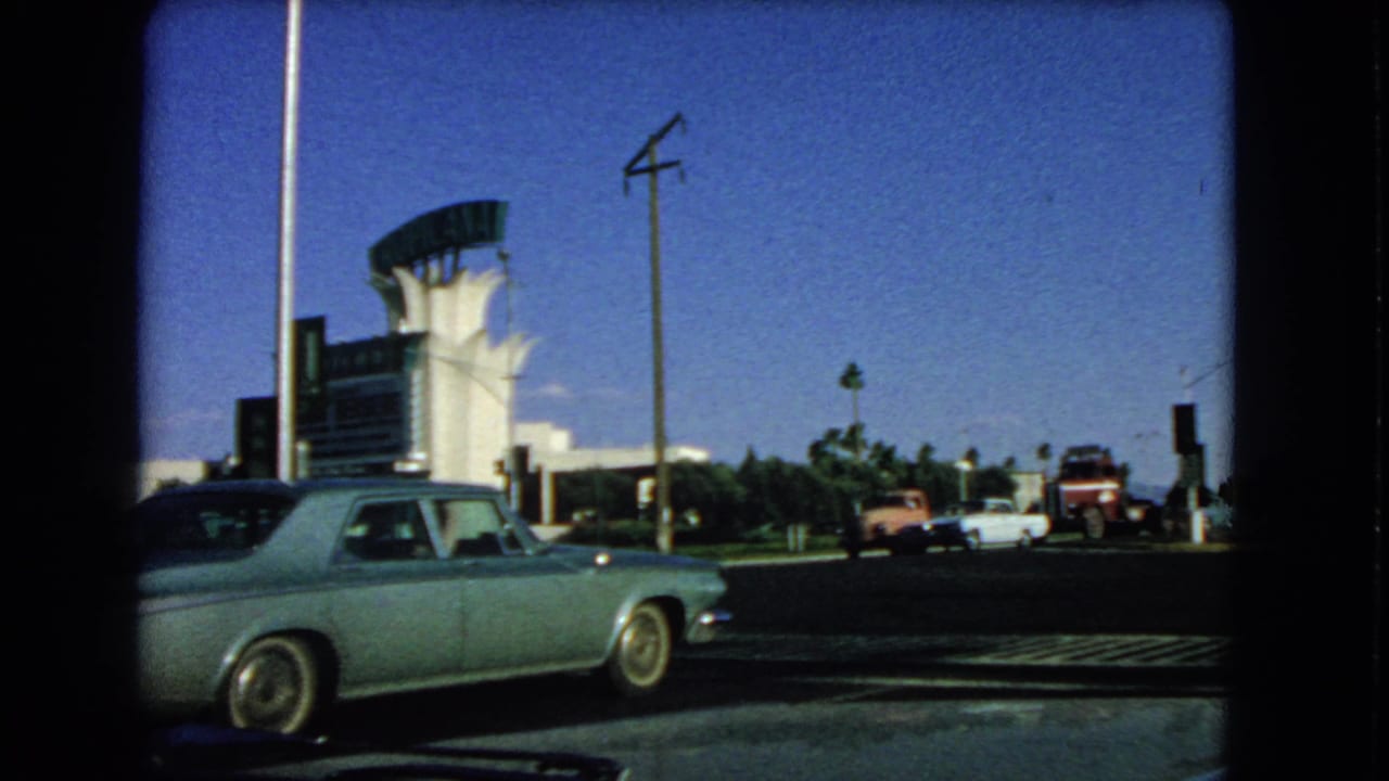 Final 1966 Las Vegas frame showing the blue sedan, distant vintage cars, roadside poles, palms, and a white building with a curved sign form.