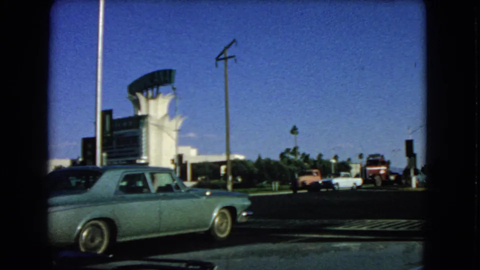 Final 1966 Las Vegas frame showing the blue sedan, distant vintage cars, roadside poles, palms, and a white building with a curved sign form.