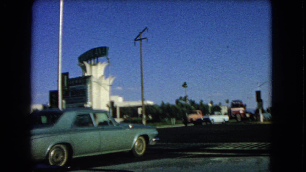 1966 Las Vegas 8mm frame showing a light blue sedan passing a sunlit intersection near a mid-century style roadside building.
