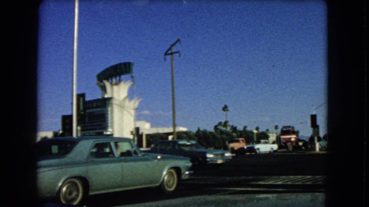 1966 Las Vegas street view with a light blue sedan, cross traffic, signals, and a distinctive mid-century roadside structure.